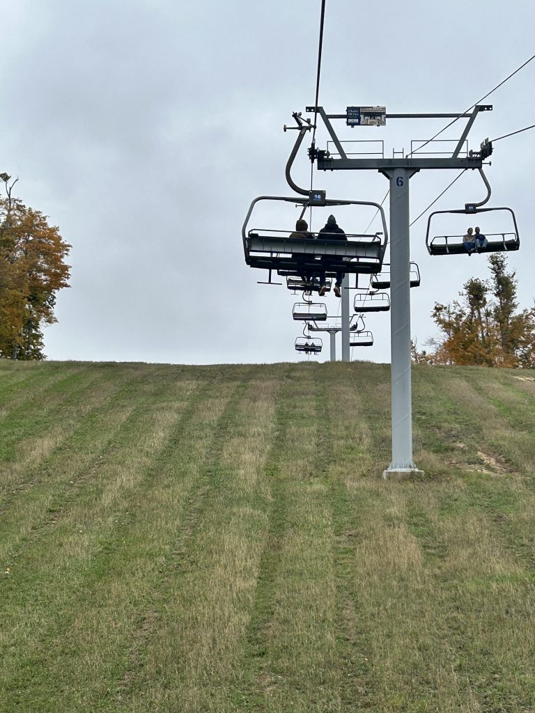 A ski lift carrying passengers up a green grassy slope under a cloudy sky, with autumn foliage in the background.