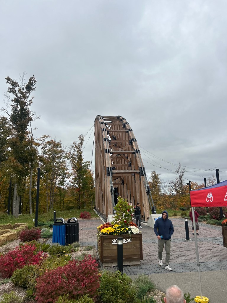A wooden bridge with a curved design, surrounded by colorful autumn foliage and flowers, with people near a sign reading 'Sky Lodge'.