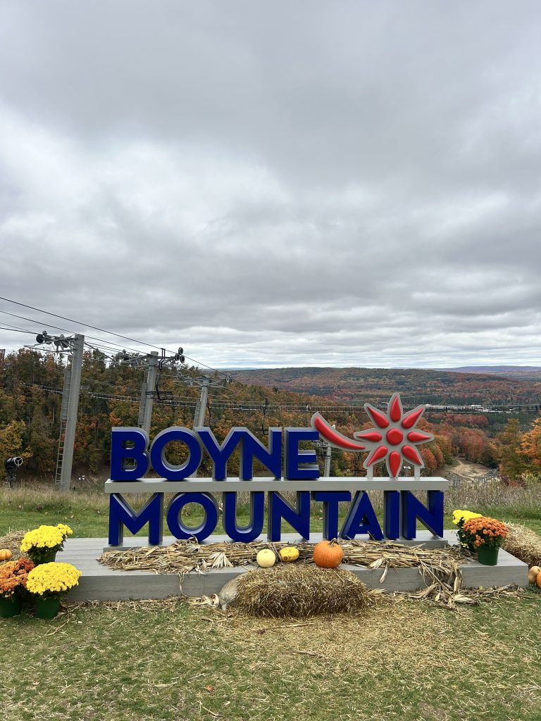 Sign for Boyne Mountain with colorful autumn foliage in the background, featuring pumpkins and flowers in front, under a cloudy sky.