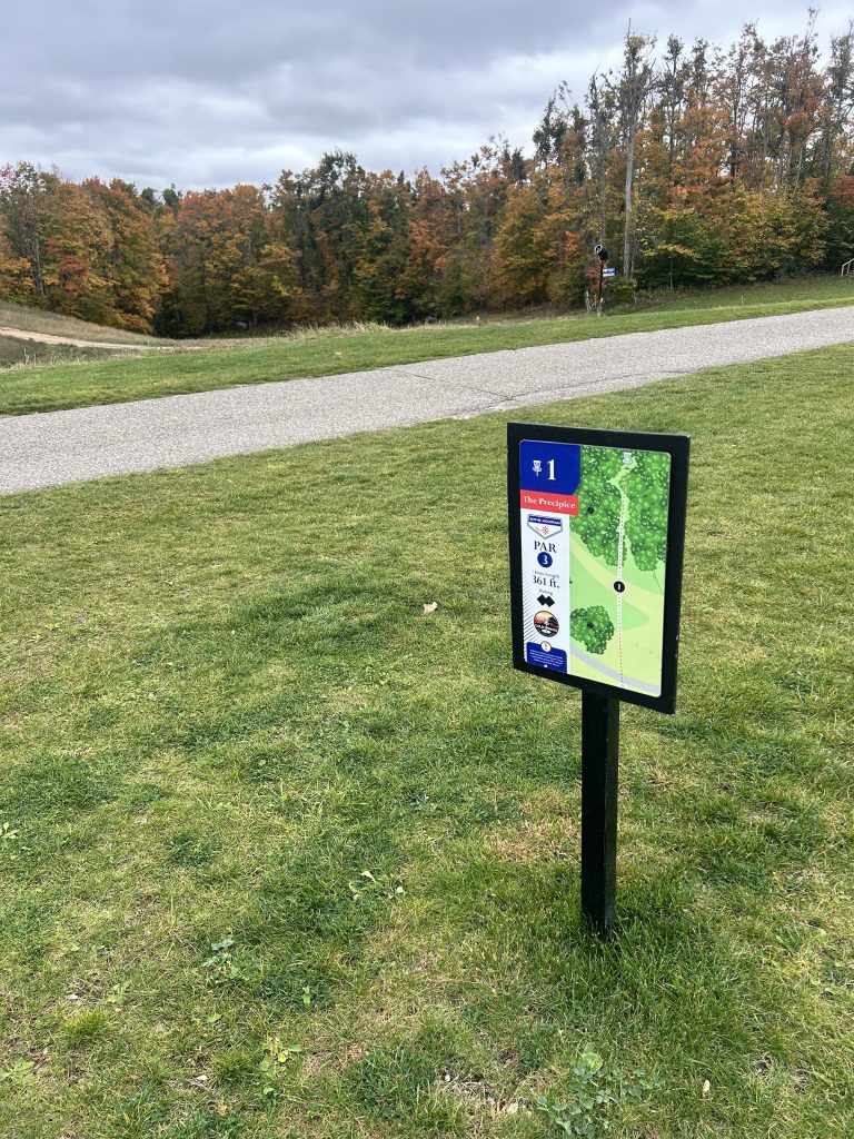 A sign marking the first hole of a disc golf course, titled 'The Precipice,' displaying details such as 'PAR 3' and a distance of '361 ft.' The background features trees with autumn foliage.