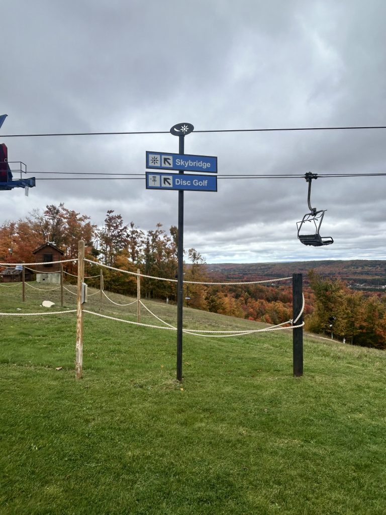 Sign directing to Skybridge and Disc Golf with a chairlift in the background amidst a mountainous landscape with fall foliage.