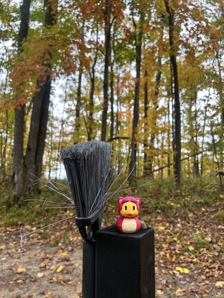 A cute toy character resembling a bear sits on top of a wooden post next to a broom with frayed bristles, surrounded by colorful autumn foliage and trees.