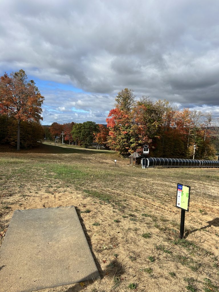 A scenic view of a ski slope in autumn, featuring vibrant orange and red foliage on trees, a concrete launch pad in the foreground, and a sign indicating a ski run. The sky is partly cloudy.