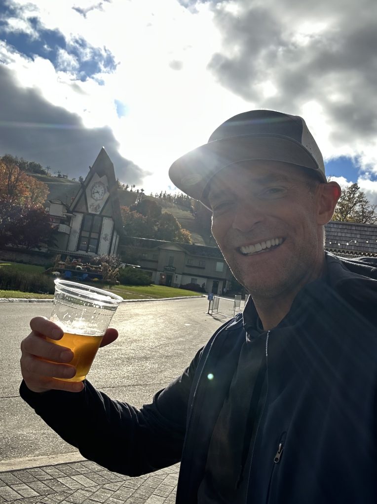 A person smiling and holding a drink in a plastic cup, with a sunlit background featuring a building and trees.